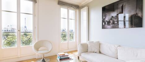 A modern living room with a white sofa, a glass coffee table, and a chair. There are large windows with a view of a cityscape.