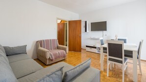 Een moderne woonkamer met een bank, een fauteuil met een plaid, een eettafel met stoelen, een aan de muur bevestigde televisie en een houten deur.