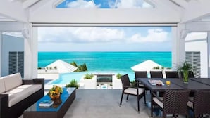 Una habitación moderna con un amplio ventanal que da a un océano turquesa. Cuenta con un rincón de comedor con mesa y sillas, además de un sofá y una mesita de centro.