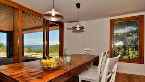 Ein Holztisch mit einer Zitronenkorb, einer Glasgießkanne und einer Tasse. Weiße Stühle und ein großes Fenster mit Blick auf Bäume und Hügel.
