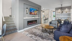 Een moderne woonkamer met een open haard, grijze muren en een eetgedeelte met een keuken op de achtergrond.