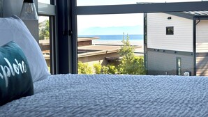 Een slaapkamer met uitzicht op de oceaan, een bed met een kussen, en een raam met uitzicht op een gebouw en bomen.