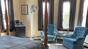 A hotel room with a large bed, two blue armchairs, a small table, a coffee maker, and a fan.