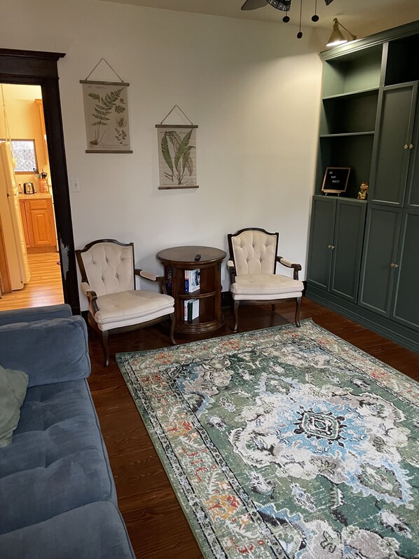 Un salón con un sofá azul, dos sillones, una mesa redonda de madera, un tapete estampado y un mueble verde con un cartelito.