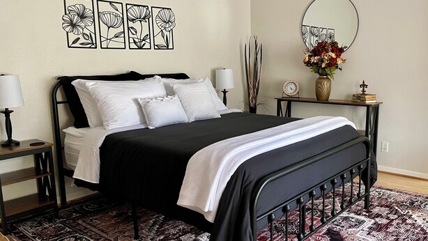 A neatly arranged bedroom with a black metal bed frame, white bedding, a patterned rug, a wall clock, a mirror, and a vase with flowers. - Mountain View Stay LLC -  The perfect place to get away, relax and enjoy nature. (Mena)
