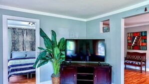 Un salon avec une grande télévision, un plancher en bois, un lit avec une literie à motifs et une grande plante verte.