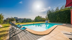 Una piscina con cubierta de madera y sillas de descanso, rodeada de follaje y un cielo despejado.