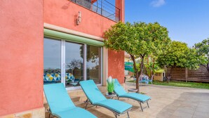 Un edificio con fachada roja, puerta de vidrio y un patio con tres sillones azules y un árbol pequeño.