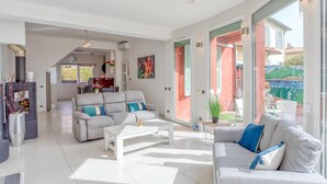 Amplia sala de estar con sofá, mesa de centro y chimenea. Se observa un comedor con mesa y sillas, y una cocina al fondo.