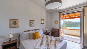 Ein Schlafzimmer mit Bett, Nachttisch, Kommode und Blick auf einen Strand durch eine offene Tür.