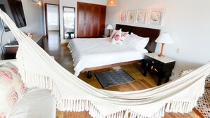 A spacious bedroom with a large bed, a hammock, wooden ceiling beams, and a view of the outside.