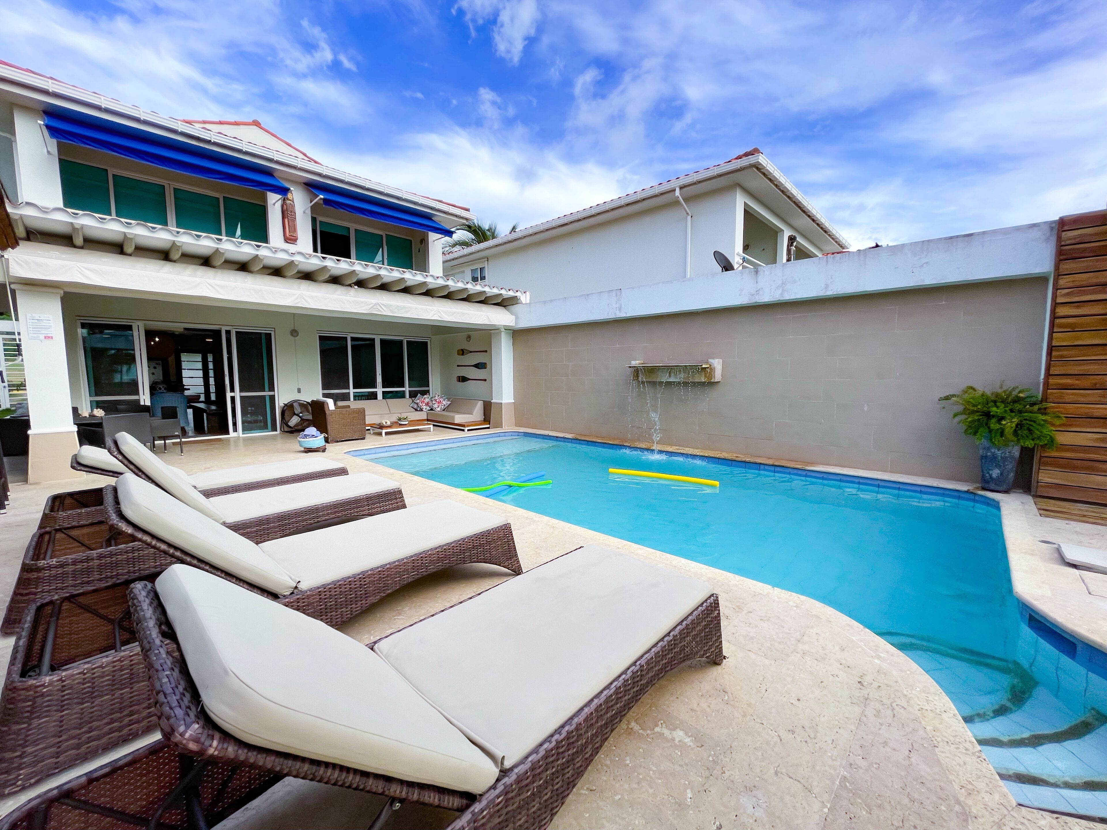 A swimming pool with lounge chairs, a covered patio, and a building with large windows and a red roof.