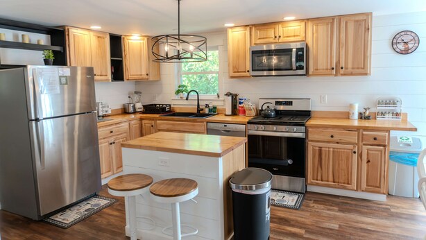 A modern kitchen with wooden cabinets, stainless steel appliances, a central island with stools, and a window providing natural light. - Deer Ridge A/C, Firepit, Pool Table, Acadia (Ellsworth)