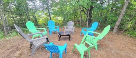 A group of Adirondack chairs arranged around a central fire pit in a forested area.