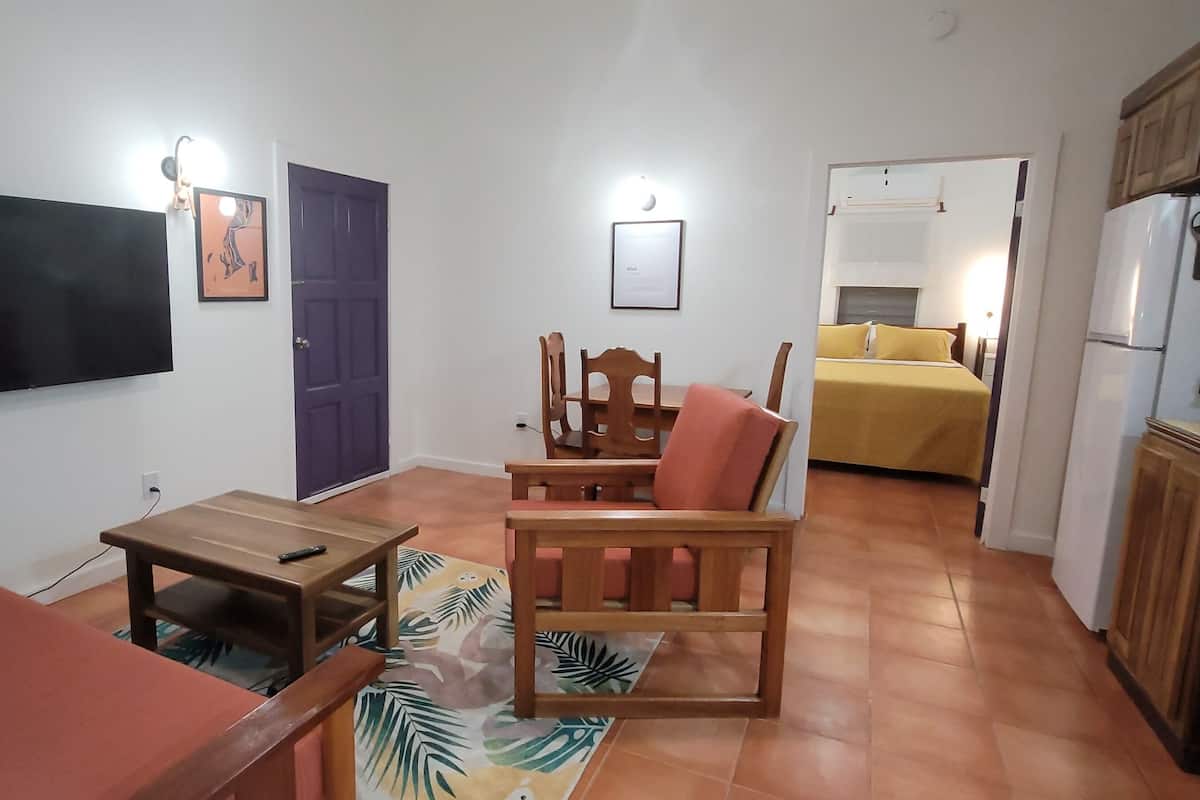 A compact living space with a flat-screen TV mounted on the wall, a small wooden table, a sofa, a chair, and a bed with a yellow bedspread.