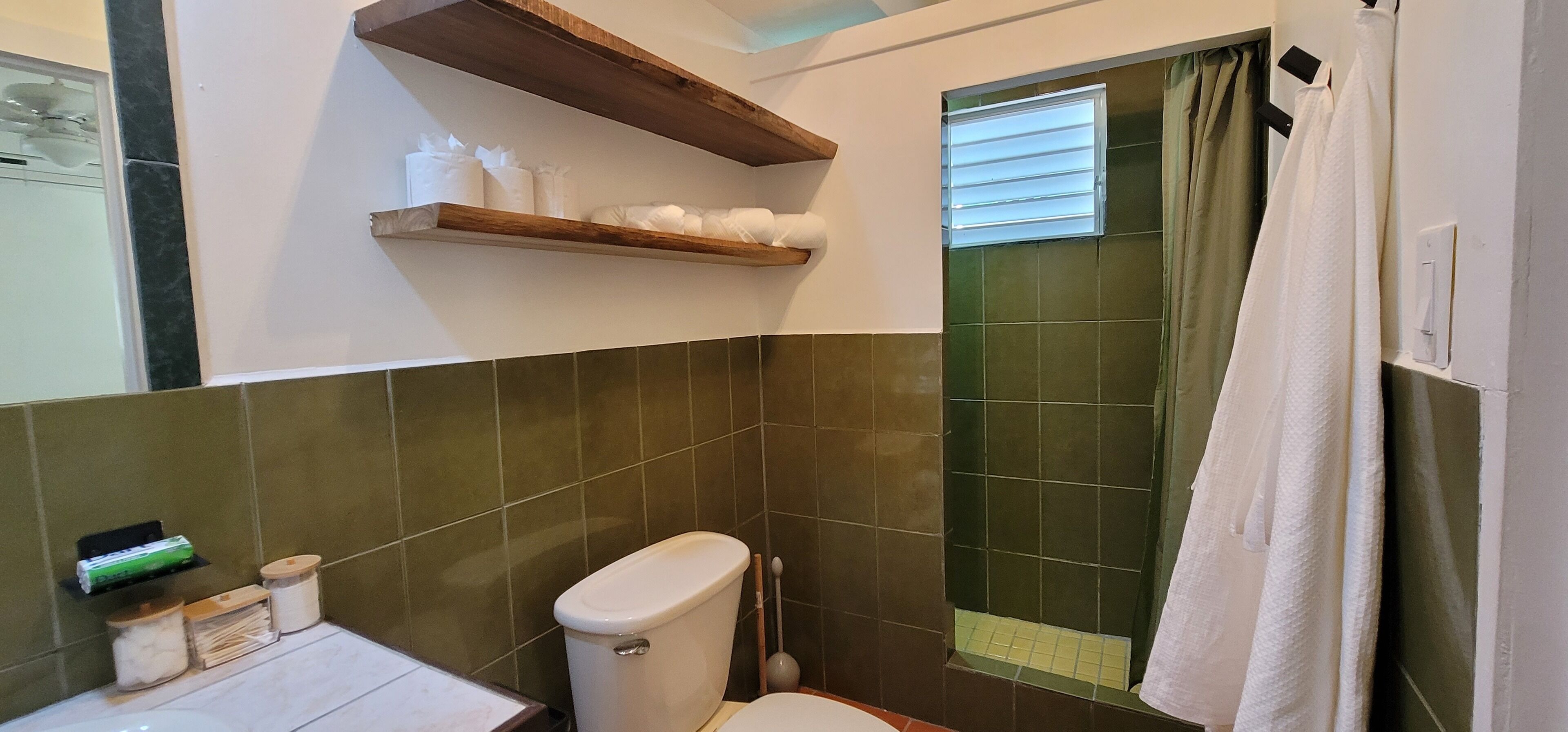 Un baño con azulejos verdes, un inodoro, un estante con artículos de aseo y una cortina de ducha.