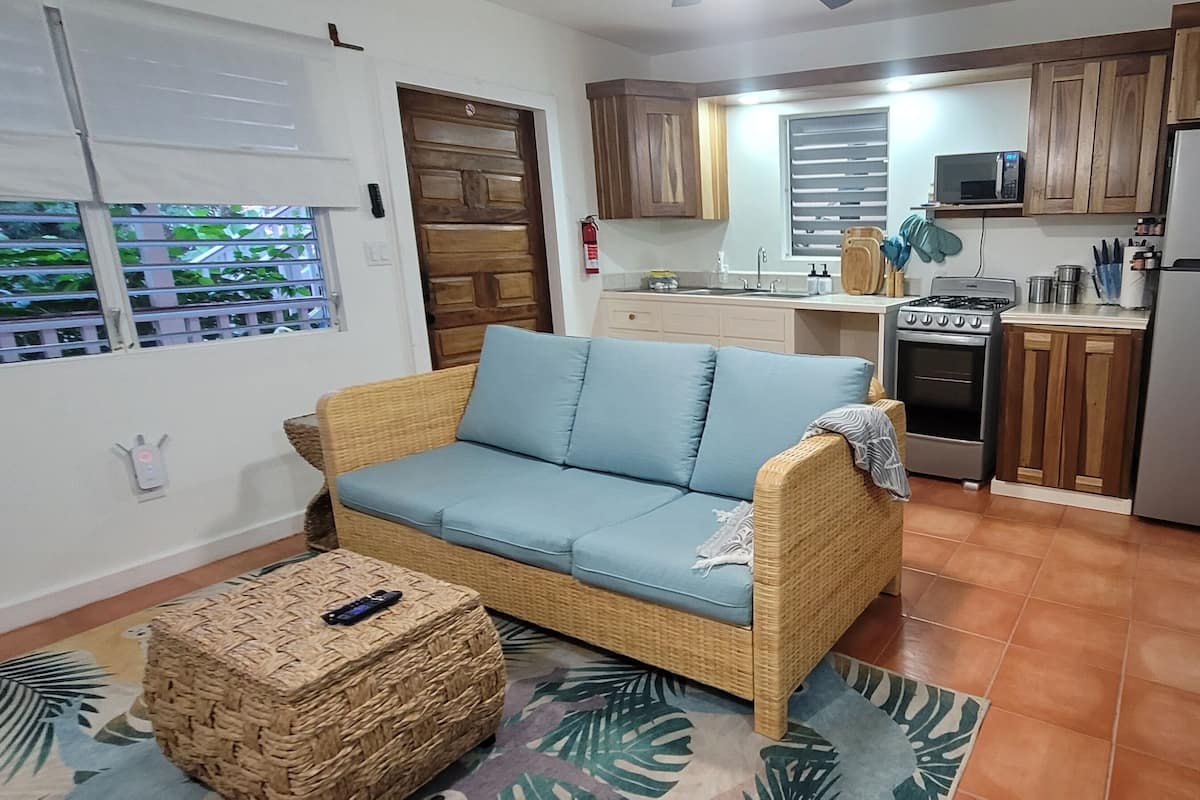 A compact living space with a sofa, a small table, a kitchen area including a stove and refrigerator, and a window with blinds.
