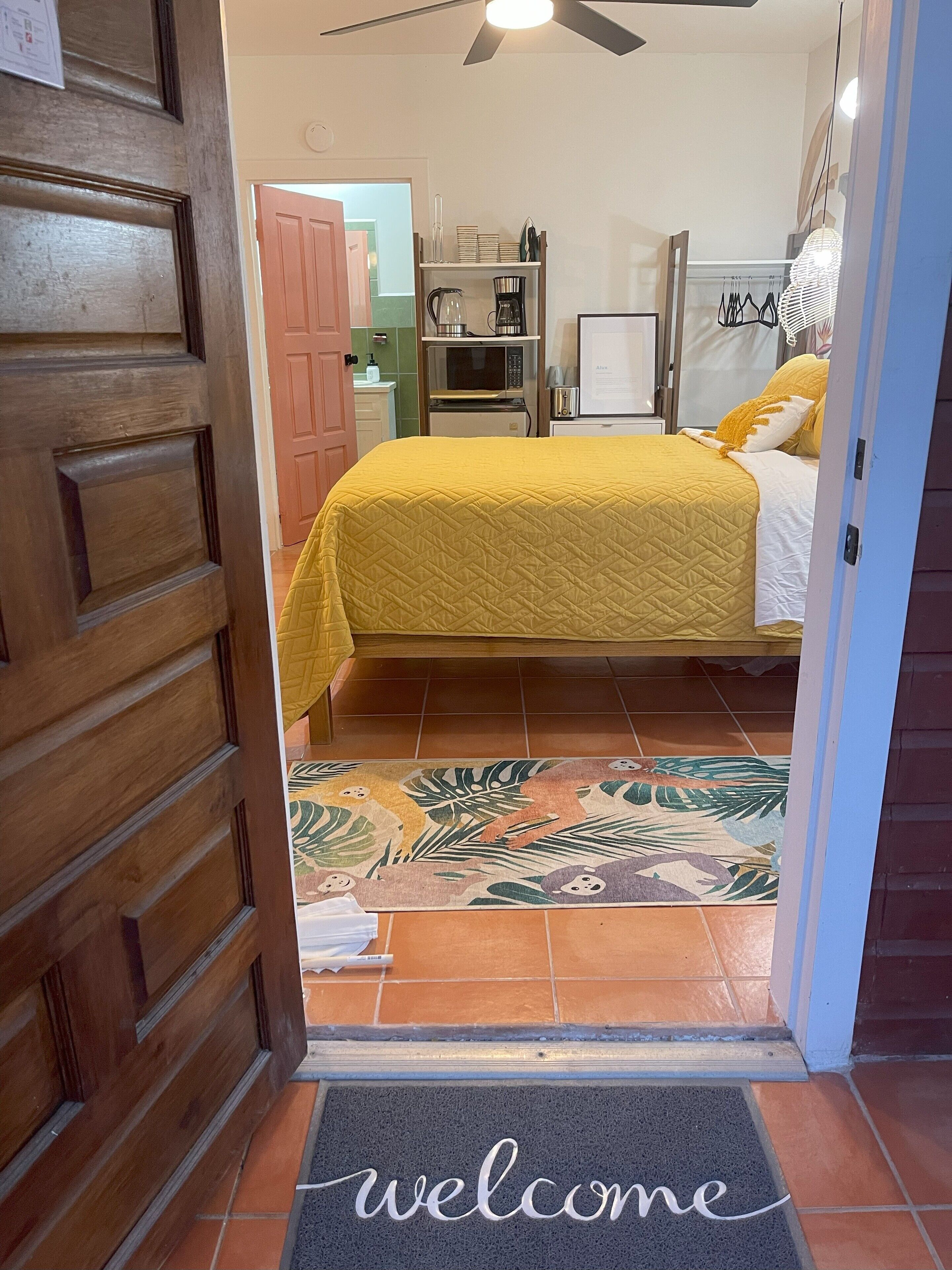 A bedroom with a bed, a wooden door, a welcome mat, and a kitchen area in the background.