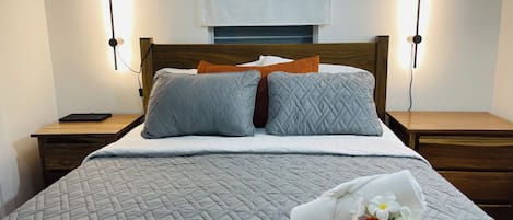 A neatly made bed with a quilted bedspread, two pillows, and a rolled towel with flowers on a wooden nightstand.