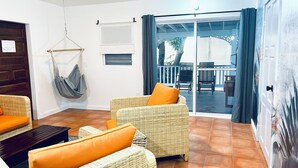 Un salón acogedor con mobiliario de mimbre, una hamaca y un ventanal corredera que da a un patio exterior.