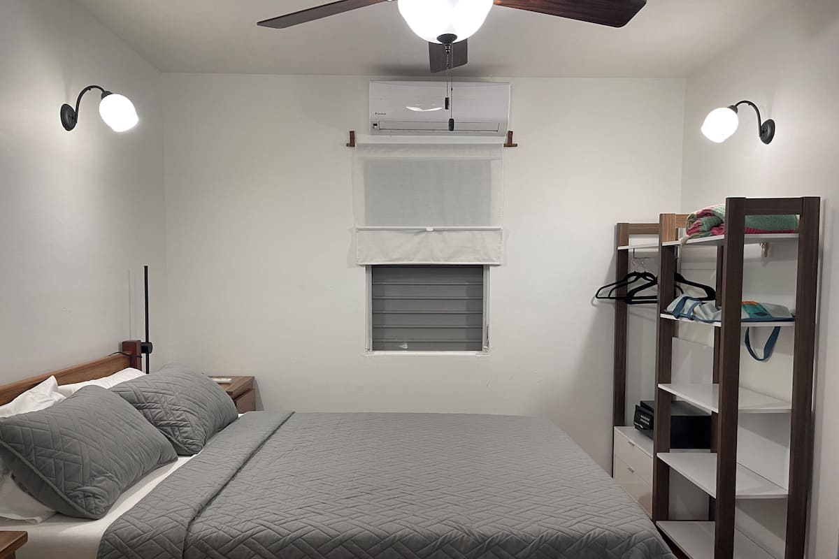 A bedroom with a bed, a wardrobe, a ceiling fan, an air conditioning unit, and a window with blinds.