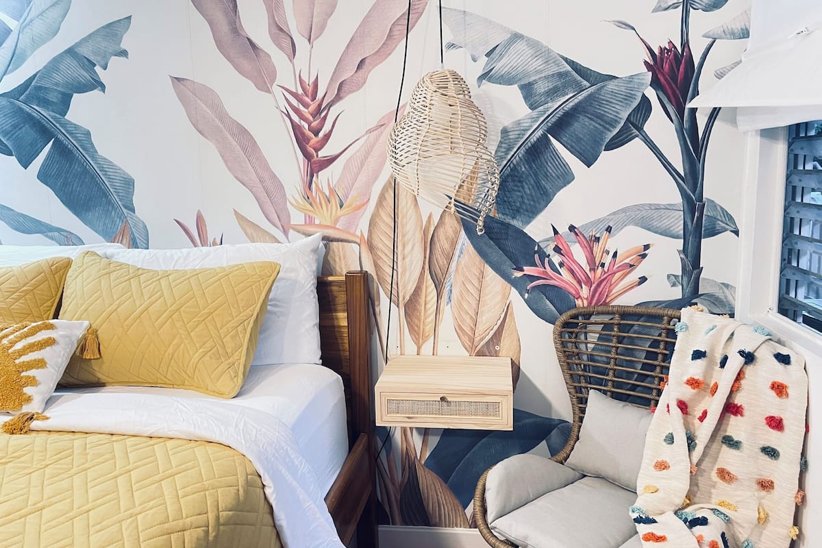 A bedroom with a bed, a chair, a wall-mounted speaker, and a large tropical-themed mural.