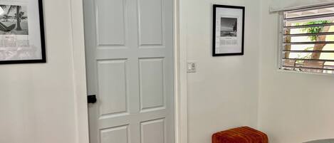 A room with a white door, a framed picture on the wall, a window with blinds, and a small red stool.