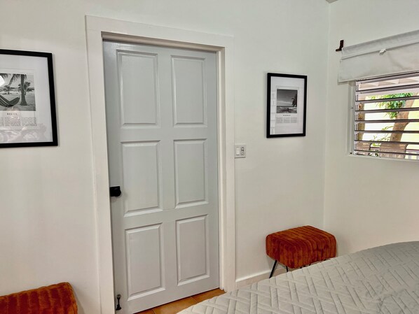A room with a white door, a framed picture on the wall, a window with blinds, and a small red stool.