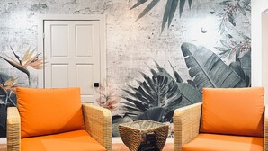 Una habitación con dos sillas de mimbre con cojines naranjas, una mesita auxiliar tejida y una puerta con un mural de pared con textura.
