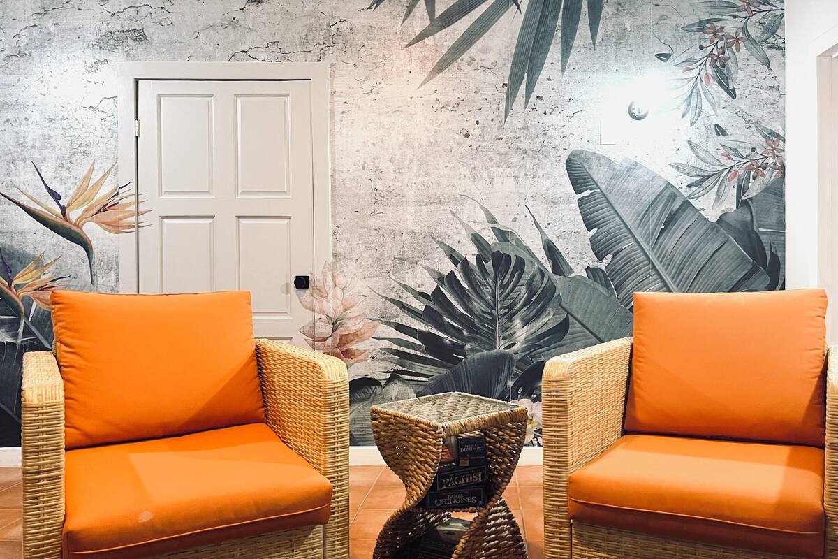 A room with two wicker chairs with orange cushions, a small woven side table, and a door with a textured wall mural.