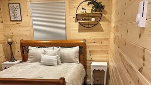 A wooden cabin-style bedroom with a bed, bedside tables, a lamp, and a wall-mounted shelf with plants.