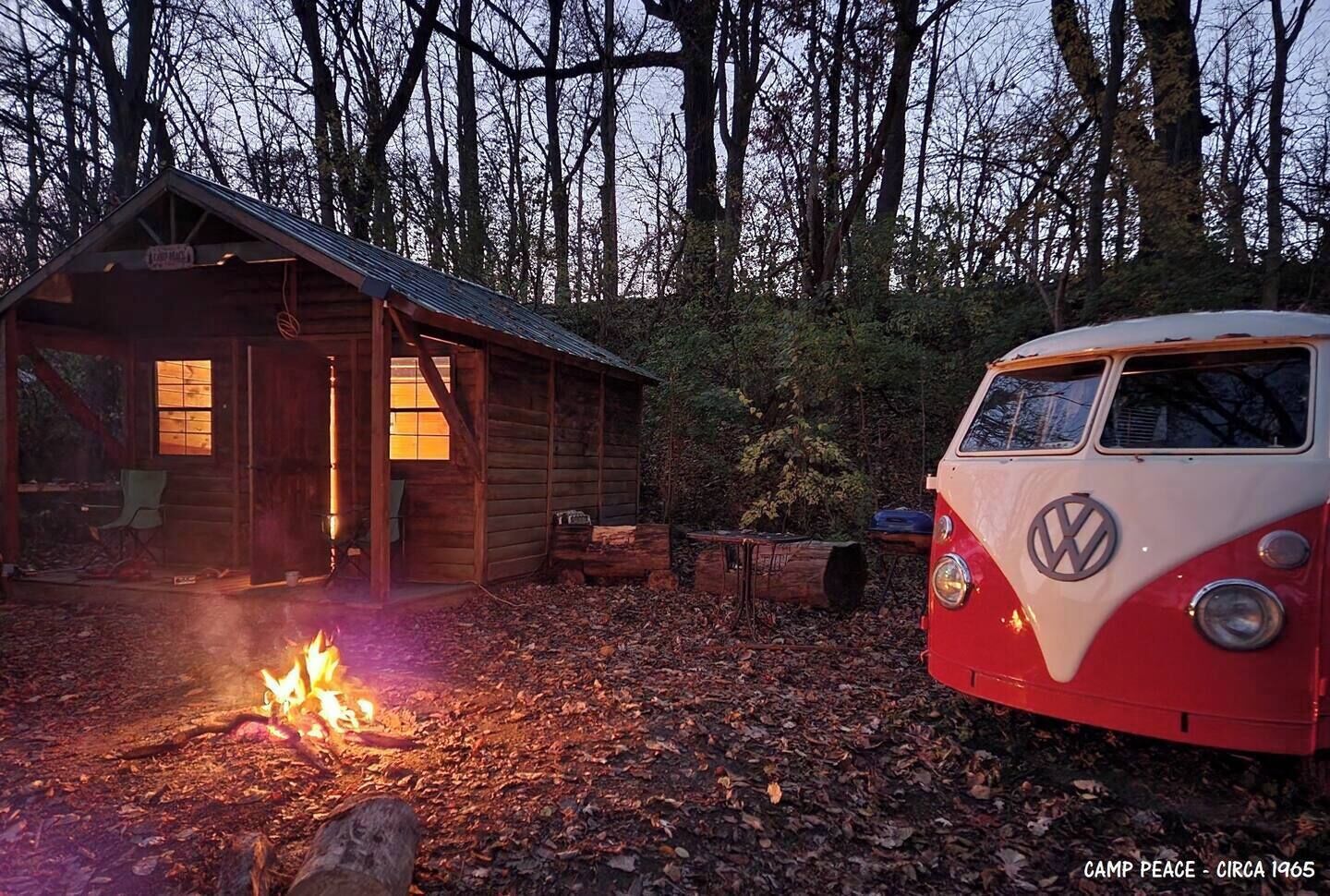 Eine Holz-Blockhütte mit Feuer davor, ein Volkswagen-Campingbus und ein bewaldeter Hintergrund.