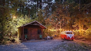 A rustic cabin with a small porch and two chairs, a vintage VW van parked nearby, surrounded by trees and string lights.