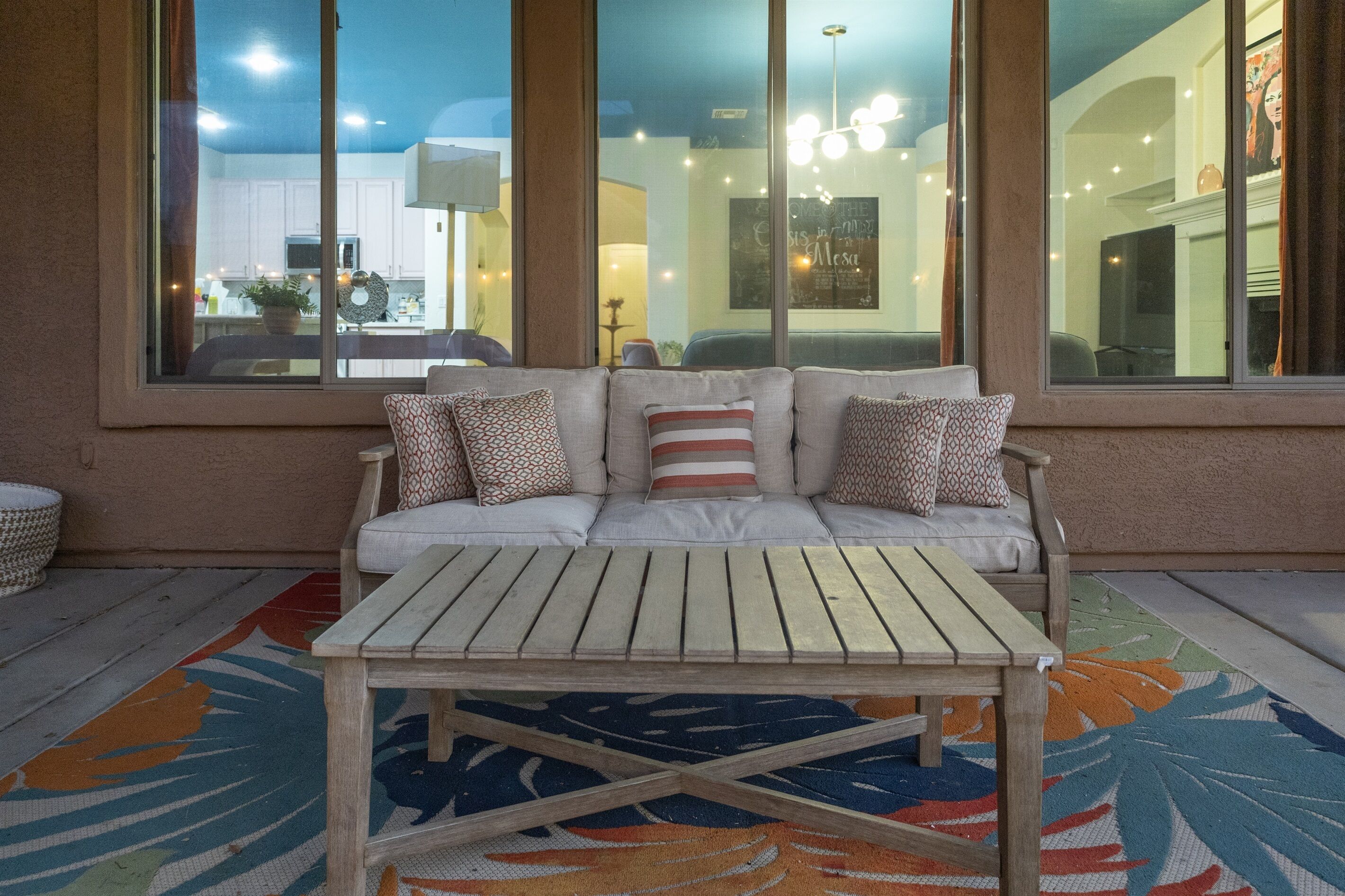 Ein gemütlicher Sitzbereich im Freien mit Holztisch und gepolstertem Sofa, große Fenster mit Blick ins Innere und einem farbenfrohen Teppich auf dem Boden.