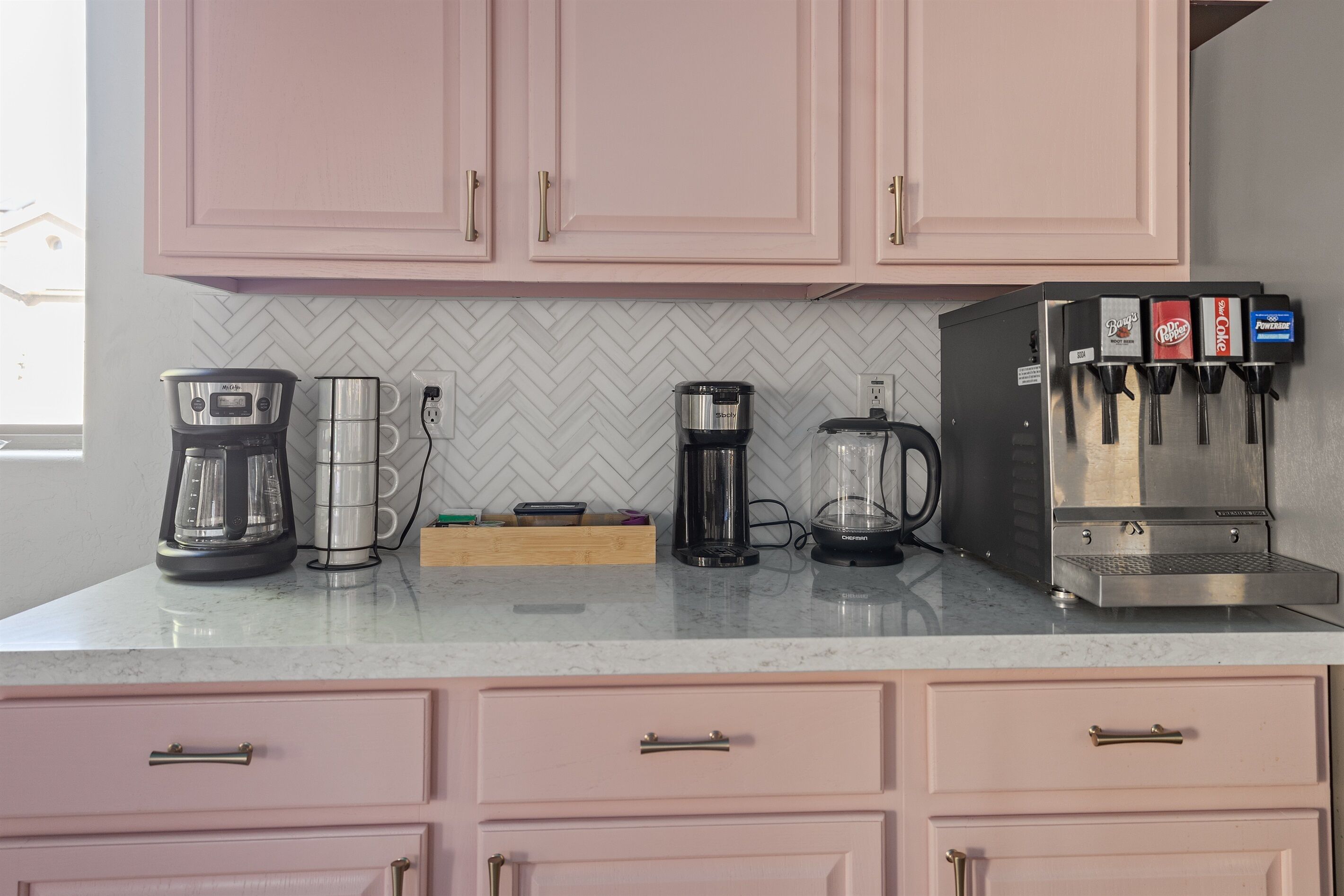 A kitchen with pink cabinets, a coffee maker, a blender, a coffee machine, and a soda dispenser.