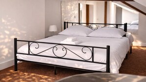 Un letto rifatto con lenzuola bianche e una struttura in metallo nero, una lampada da comodino e vista su una stanza con tetto spiovente e travi in legno.