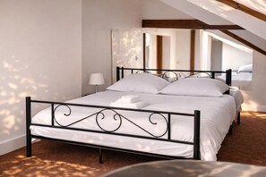 Un letto rifatto con lenzuola bianche e una struttura in metallo nero, una lampada da comodino e vista su una stanza con tetto spiovente e travi in legno.
