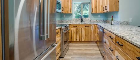 A kitchen with wooden cabinets, a stainless steel refrigerator, a microwave, and a window with a view of trees.