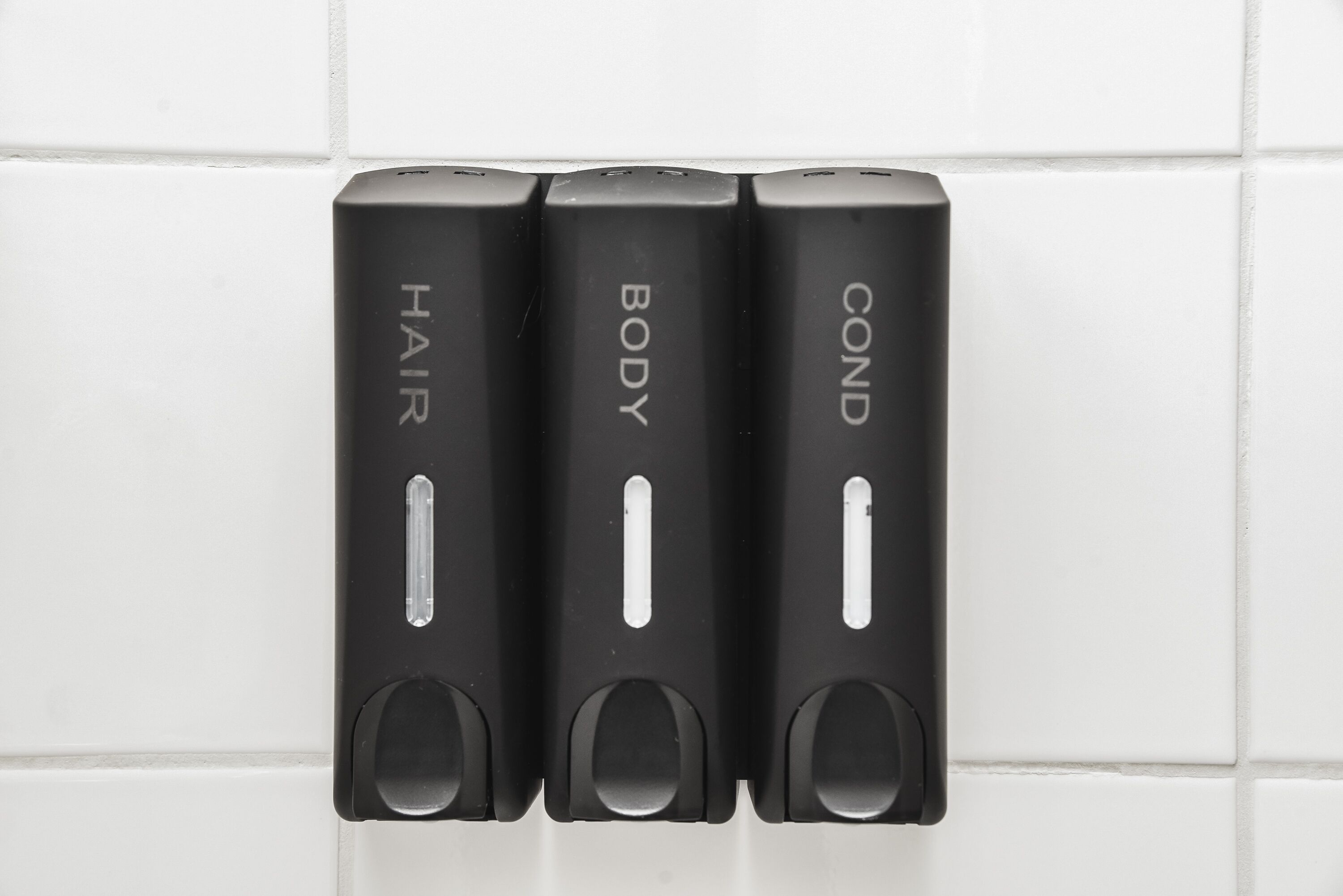 Three black dispensers labeled "HAIR," "BODY," and "COND" on a white tiled wall.