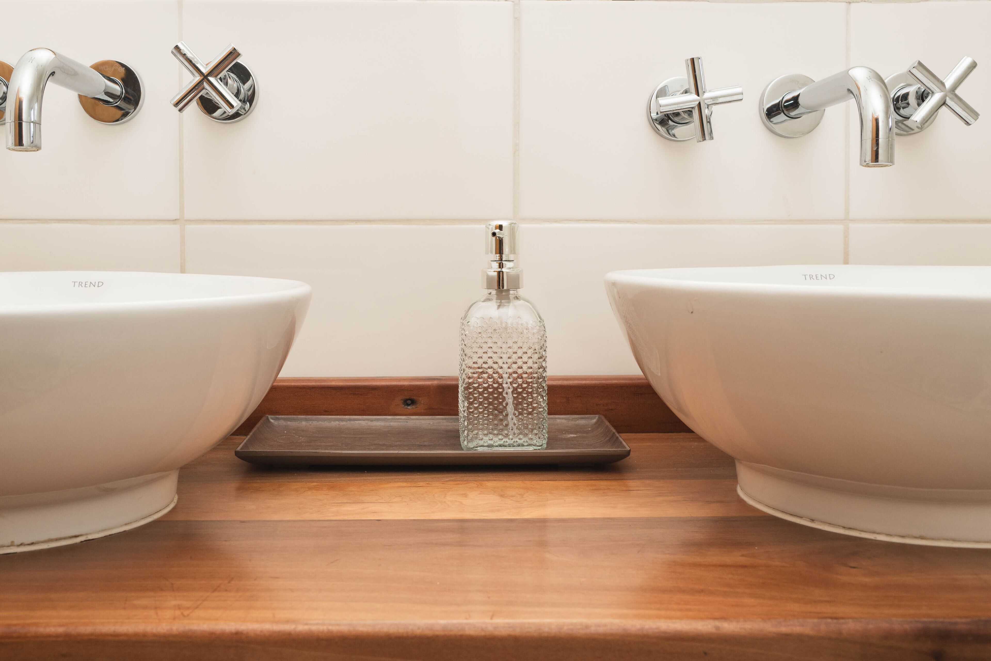 Two white sinks with chrome faucets and a clear glass bottle on a wooden tray between them.