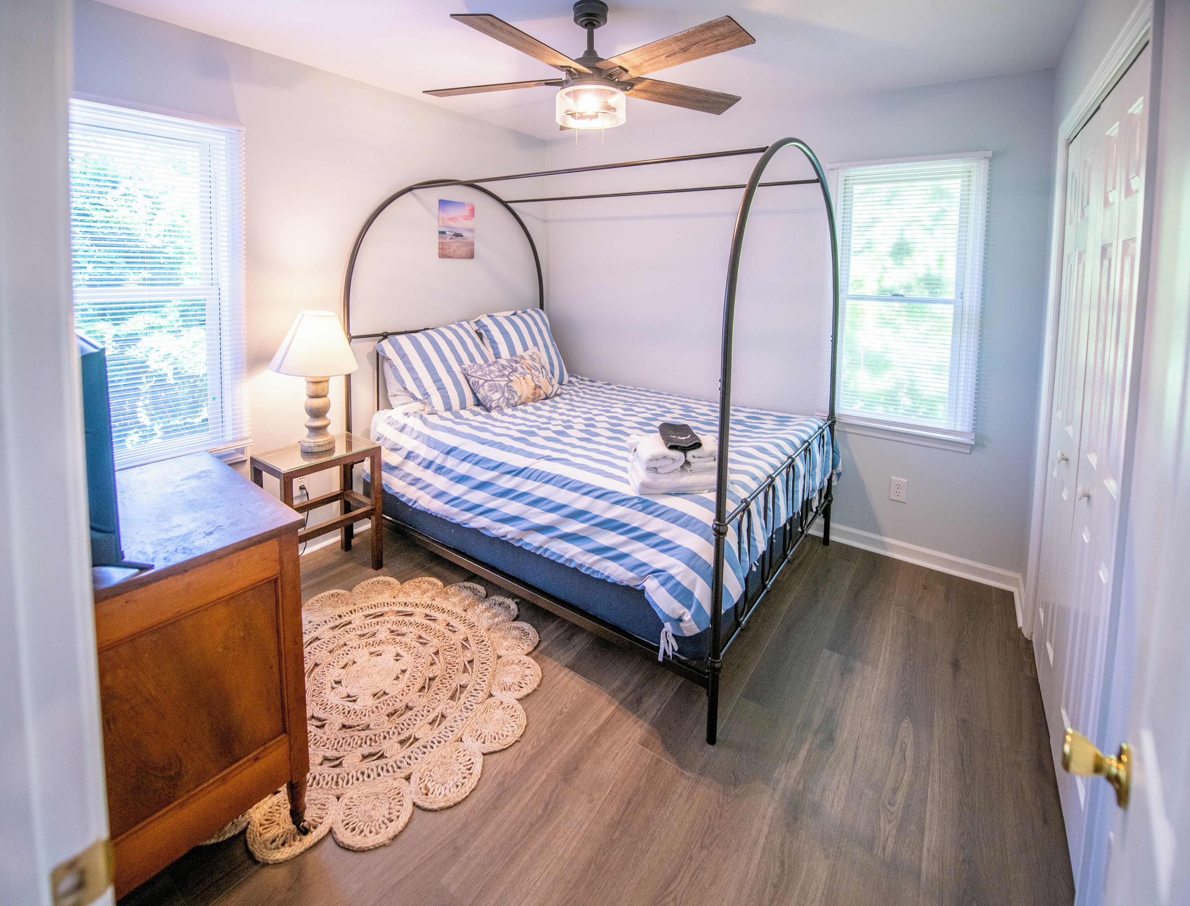 A bedroom with a four-poster bed, a bedside table with a lamp, a woven rug, and a ceiling fan.