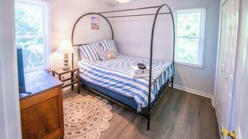 A bedroom with a four-poster bed, a bedside table with a lamp, a woven rug, and a ceiling fan.