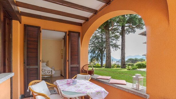 Eine Terrasse mit Tisch und Stühlen, ein Korbsessel, ein Bett mit weißem Kopfteil und Blick auf einen Garten und Berge.