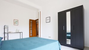 Une chambre à coucher comprenant un lit, une armoire, une petite table avec quelques objets, et une porte donnant sur une autre pièce.