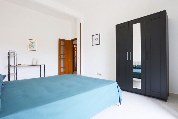 A bedroom with a bed, a wardrobe, a small table with items, and a door leading to another room.