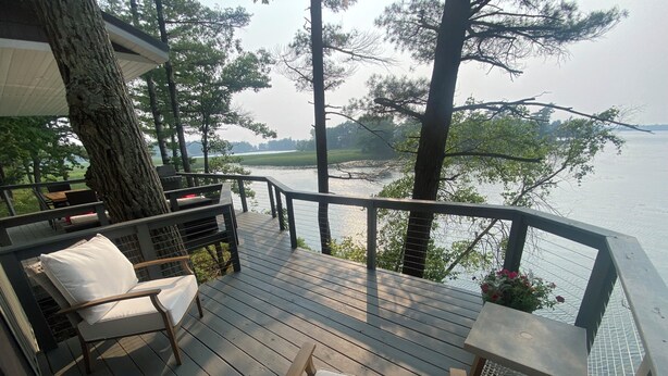 Une terrasse en bois, agréée de mobilier d’extérieur, notamment des chaises et une table, qui offre une vue sur une étendue d’eau et est entourée d’arbres.