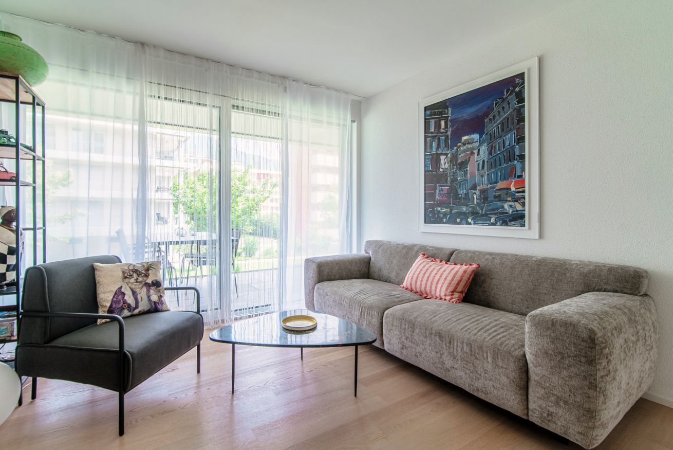 Ein Wohnzimmer mit grauem Sofa, einem schwarzen Sessel, einem Glastisch und einem Stadtbild an der Wand.