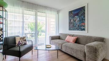 Ein Wohnzimmer mit grauem Sofa, einem schwarzen Sessel, einem Glastisch und einem Stadtbild an der Wand.