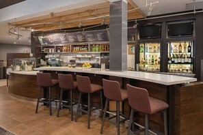 Zona de bar moderna con barra curva, taburetes, panel de menú y estantes con botellas.