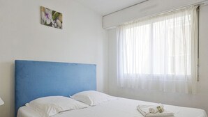 A neatly made bed with white linens and a blue headboard, a window with sheer curtains, and a small painting on the wall. - Appartement Herta - Welkeys (Biarritz)
